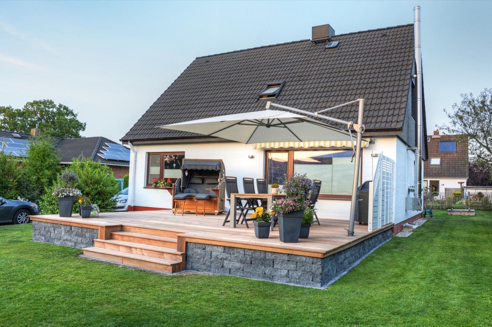 Holzterrasse mit Treppe, Esstisch und Sonnenschirm am weißen Einfamilienhaus mit grünem Rasen